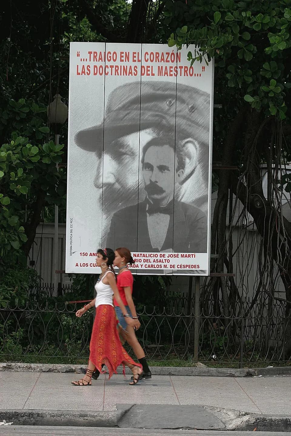 José Martí - Havana, Cuba. 2 young women walk in front of a poster showing the faces of Fidel Castro and José Martí with the quote "I carry the doctrines of the master in my heart", to suggest that Ca