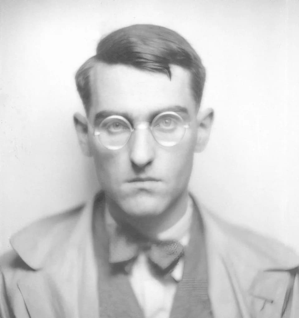 Jean Anouilh - Jean Anouilh fixe l'objectif sans émotion particulière. Il porte des lunettes rondes cerclées de métal. Il est vêtu d'un pardessus mastic, d'un veston gris, d'une chemise claire et d'un