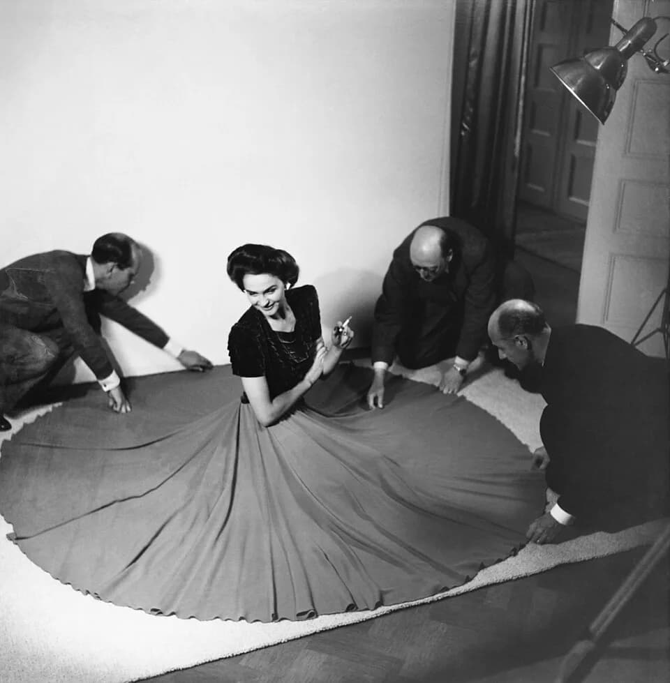 David Niven - Modellen Hjördis Genberg fotograferas för Stil. Hon sitter på en vit matta, klädd i mörk överdel och vid kjol med en cigarett i handen. Tre män, fr. v. herr Paulsen, herr Hilgardt och ka