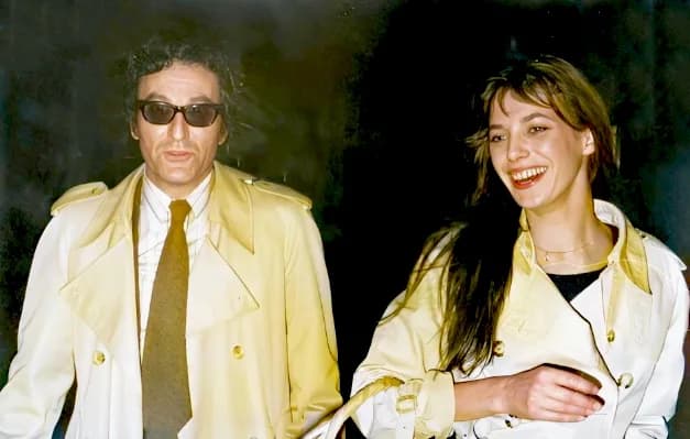 John Barry - Un couple, la nuit, photographiés au flash. Lui, à gauche, est un homme d'une quarantaine d'années, face caméra, cheveux bruns ondulés et portant des lunettes noires, un pardessus mastic 