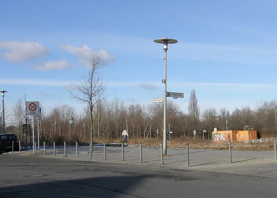 Kurt Weill - Berlin-Hellersdorf. Blick von einem Straßenknick der Janusz-Korczak-Straße, wo von links die Senftenberger Straße einmündet, zum Kurt-Weill-Platz. Dieser ist die noch wilde Fläche voraus,