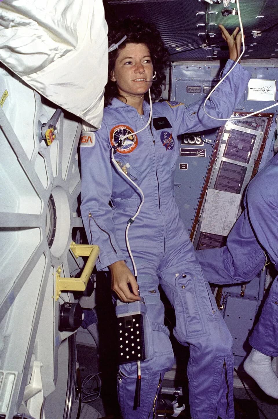 Sally Ride - On Challenger's middeck, Mission Specialist (MS) Sally Ride, wearing light blue flight coveralls and communications headset, floats alongside the middeck airlock hatch.