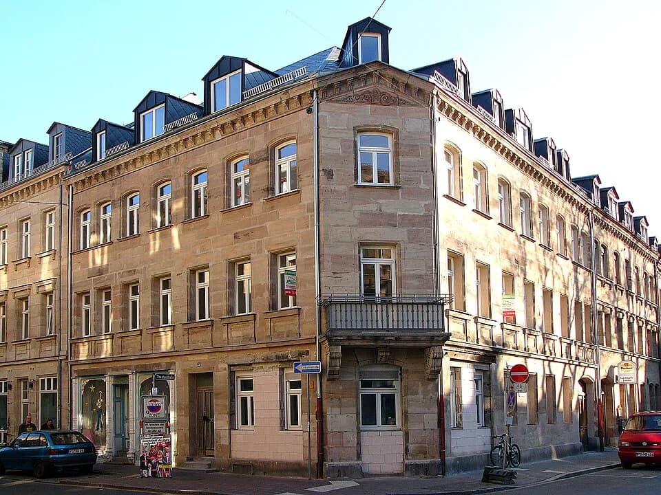 Henry Kissinger - Mietshaus in Ecklage; dreigeschossiger Satteldachbau mit Sandsteinfassade, Flachgiebel und Eisenbalkon an der abgeschrägten Ecke, Konsoltraufgesims und Neurenaissance-Ladenstock aus