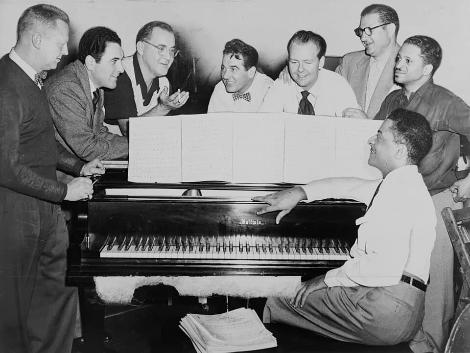 Benny Goodman - Benny Goodman (third from left) with some of his former musicians, seated around piano. 
Left to right: Vernon Brown, George Auld, Gene Krupa, Clint Neagley, Ziggy Elman, Israel Crosby