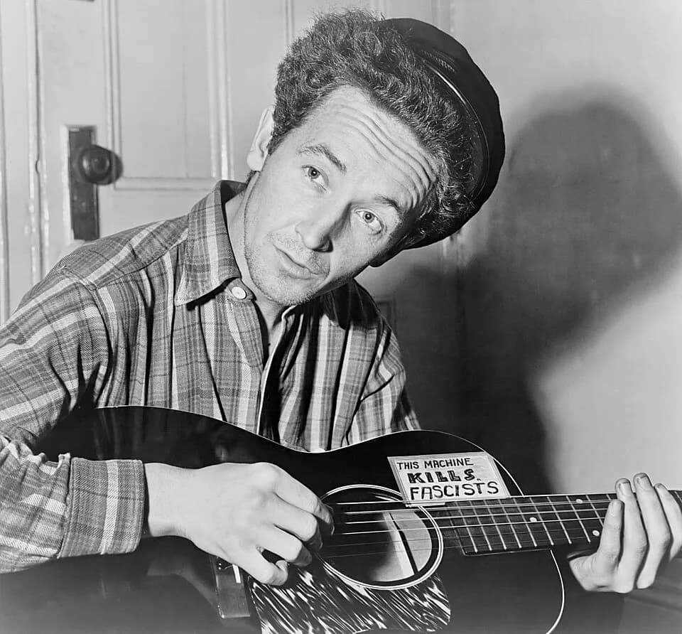 Woody Guthrie - Title: Woody Guthrie, half-length portrait, seated, facing front, playing a guitar that has a sticker attached reading: This Machine Kills Fascists