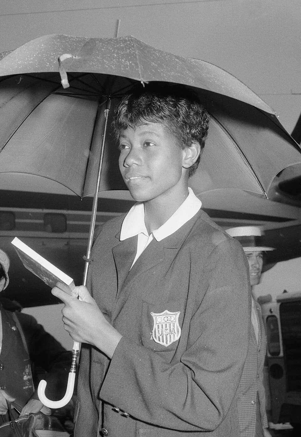 Wilma Rudolph - Aankomst Amerikaanse Olympische atleten op Schiphol, W. Rudolph poseert onder paraplu voor de fotografen