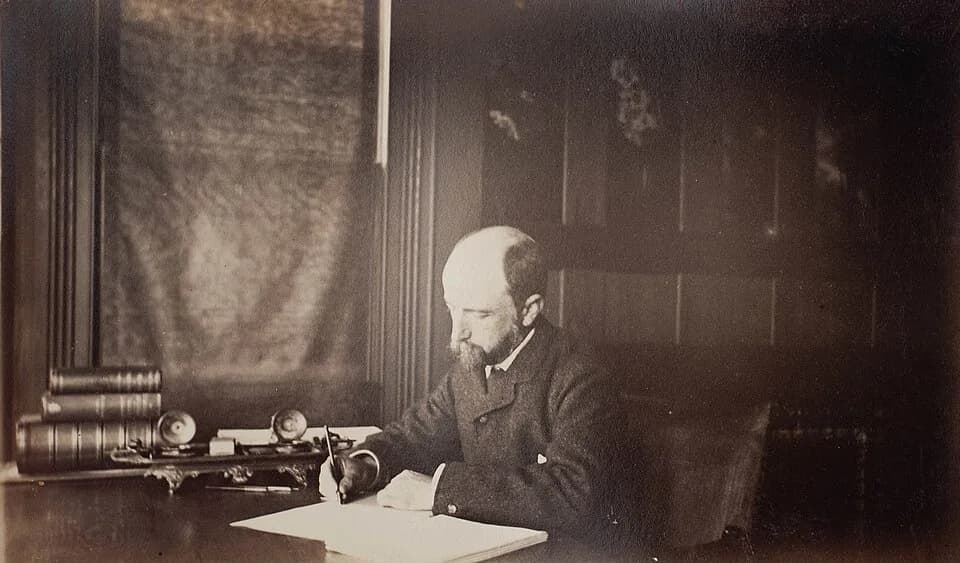 Henry Adams - Henry Adams seated at desk in dark coat, writing. Photograph by Marian Hooper Adams, 1883. Album page: 17.4 cm. x 25.5 cm.; photograph: 11.5 cm x 20 cm. From the Marian Hooper Adams phot