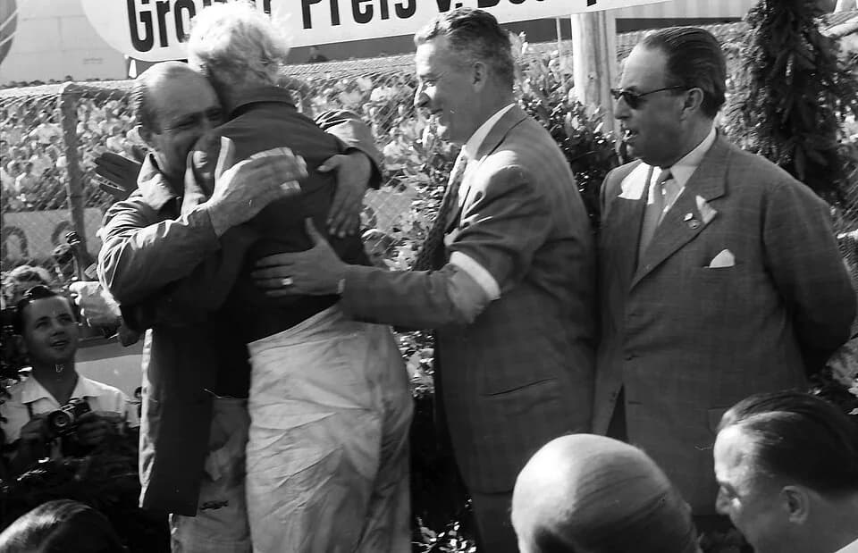 Mike Hawthorn - Second-placed driver, Mike Hawthorn (back to camera), embraces the race winner, Juan Manuel Fangio (left), on the podium for the 1957 German Grand Prix.